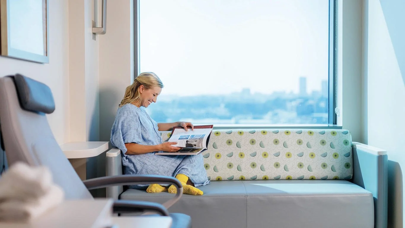 A woman in a hospital gown sitting on a medical examination room bench, reading a magazine, with a hospital bed partially visible in the foreground and a large window showing a cityscape in the background.