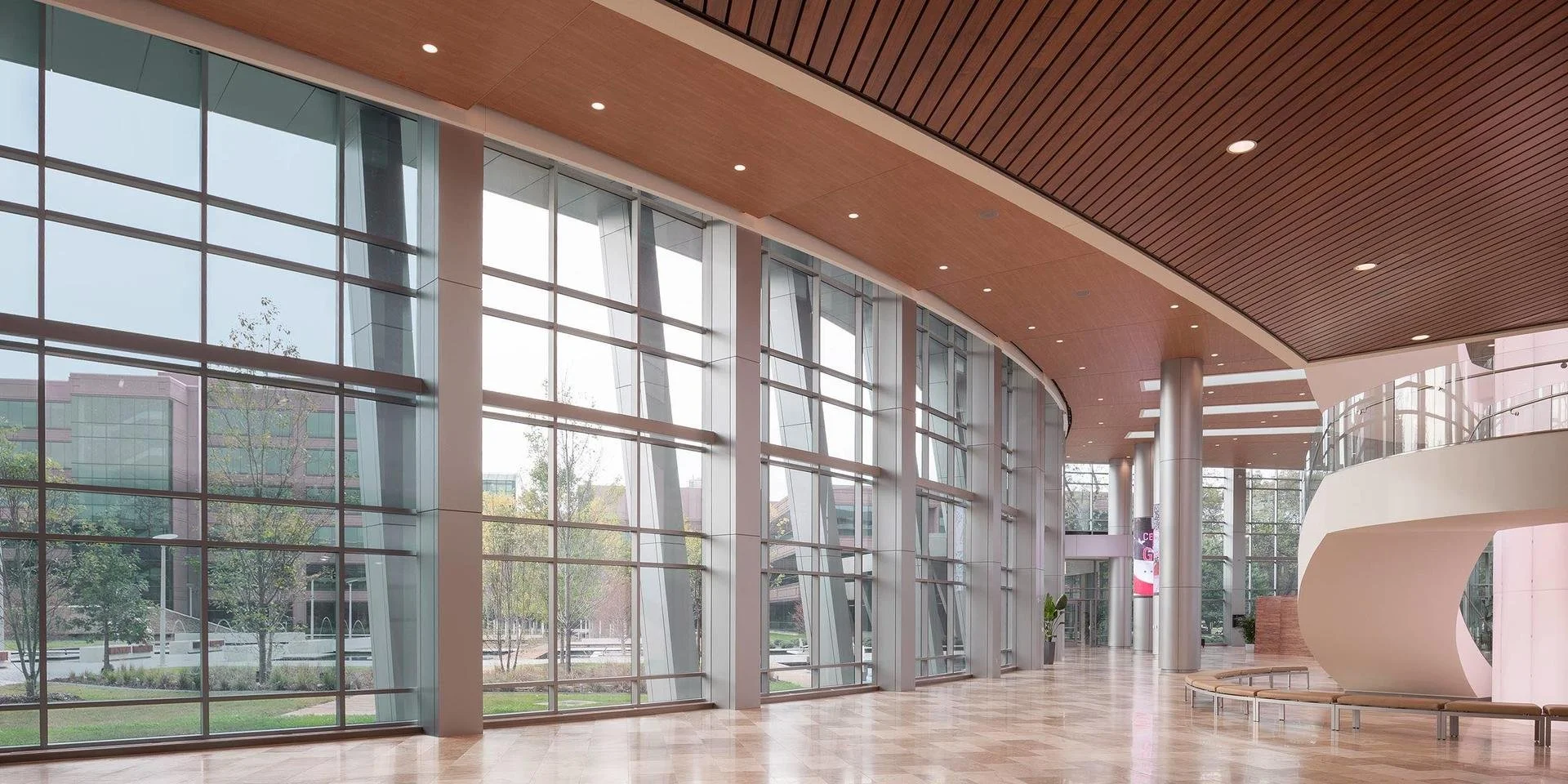 Modern, spacious indoor area with large glass windows, wooden ceiling, metal columns, and a curved white bench.