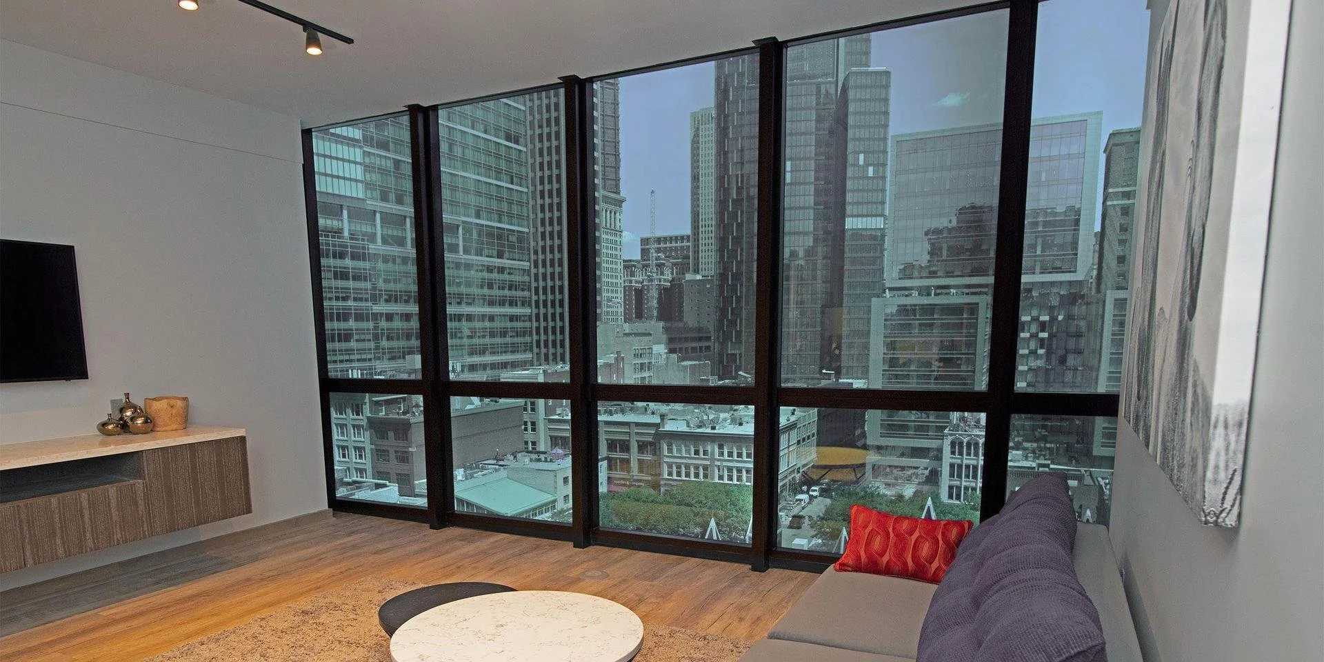 Interior view of a modern high-rise apartment living room with large floor-to-ceiling windows showing a city skyline of tall office buildings. The room has a purple sofa with a red patterned cushion, a white marble coffee table, a wooden sideboard with decorative items, and a wall-mounted flat-screen TV.