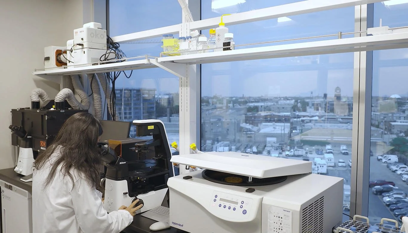 A scientist working in a laboratory using a microscope near a large window overlooking a city with buildings and a parking lot.