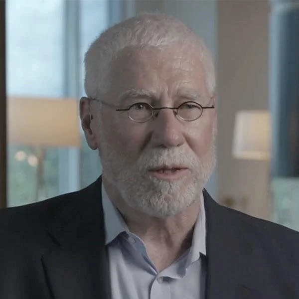 A man with white hair and beard, wearing glasses, a dark blazer, and a light shirt, sitting indoors near windows with blurred background of lamps and windows.