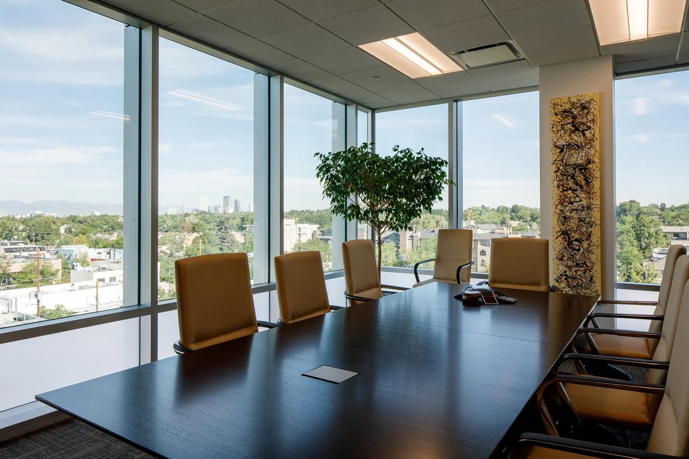 Empty conference room with a long dark table, beige chairs, a potted plant, and large windows showing city views.