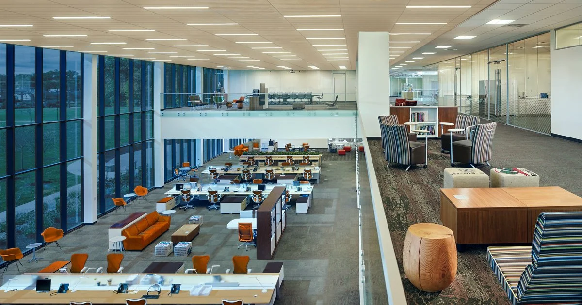 A spacious modern office with a large glass window wall, various work desks, chairs, and lounge seating in a two-story open layout.