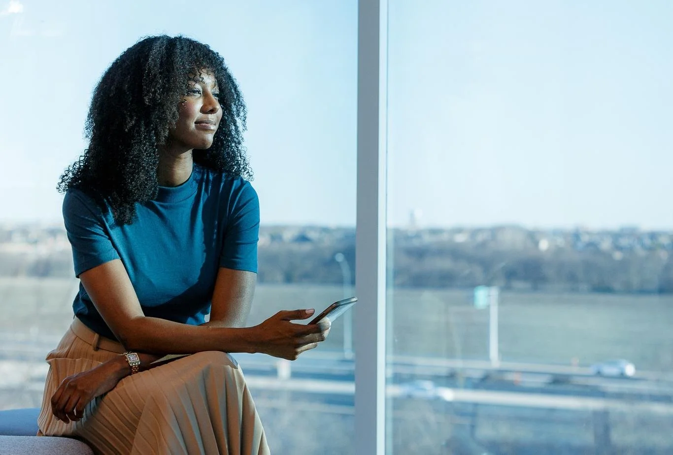 A woman with curly black hair sitting by a large window, holding a smartphone, looking thoughtfully outside on a bright day.