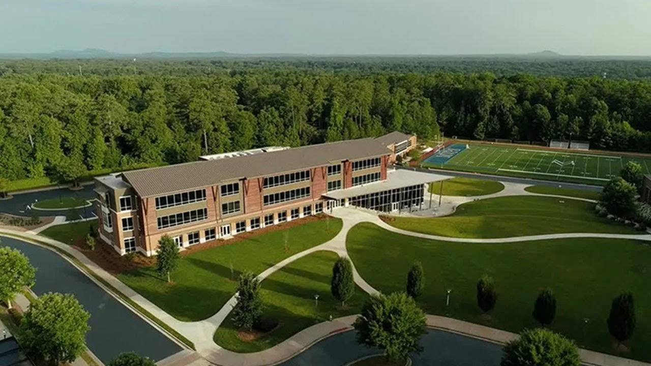 An aerial view of a large brick school building with multiple floors, surrounded by green lawns, walking paths, and a wooded area. There is a football field and a track nearby.