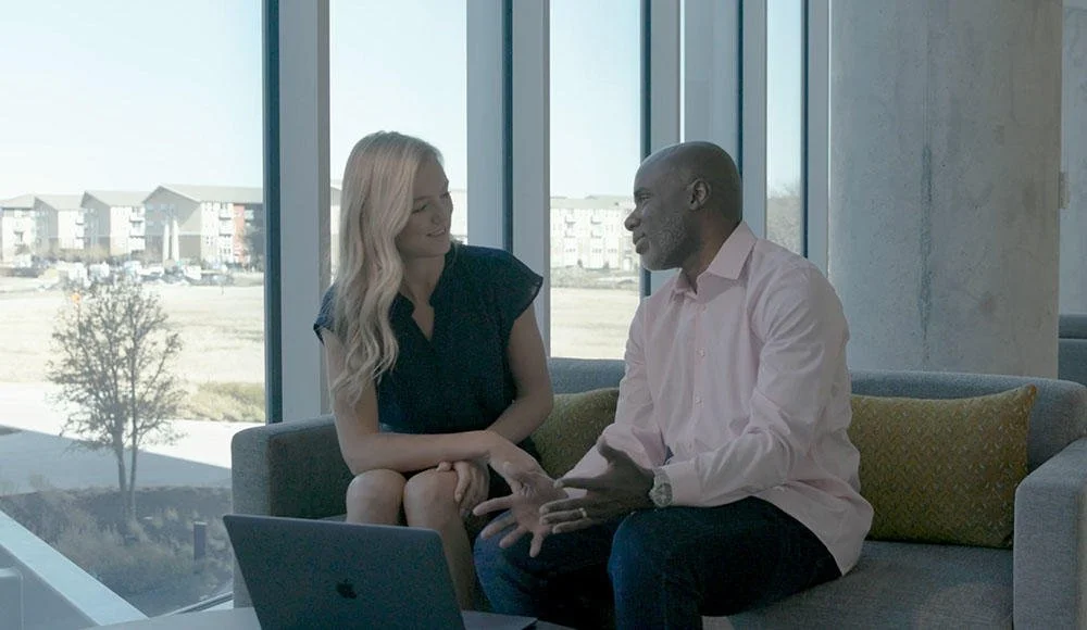 A man and a woman sitting on a sofa having a conversation in a bright room with large windows showing a parking lot and buildings outside.