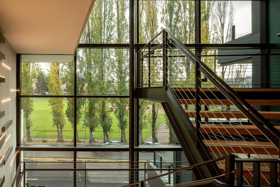Interior view of a modern building with large glass windows showing green trees and grass outside, staircase with metal railing and wooden steps.