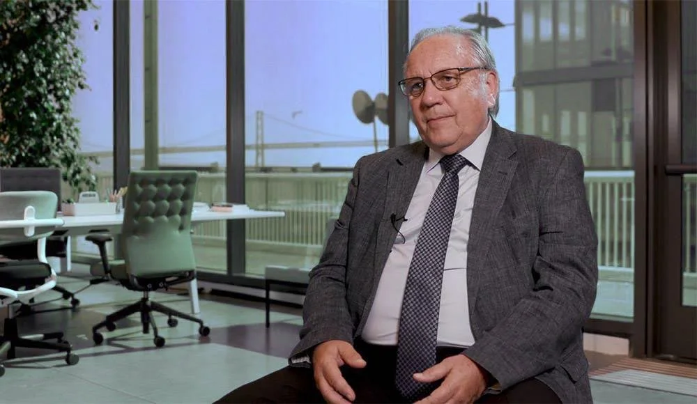 An elderly man with glasses, wearing a gray suit and patterned tie, sitting in an office conference room with large windows and a cityscape view in the background.