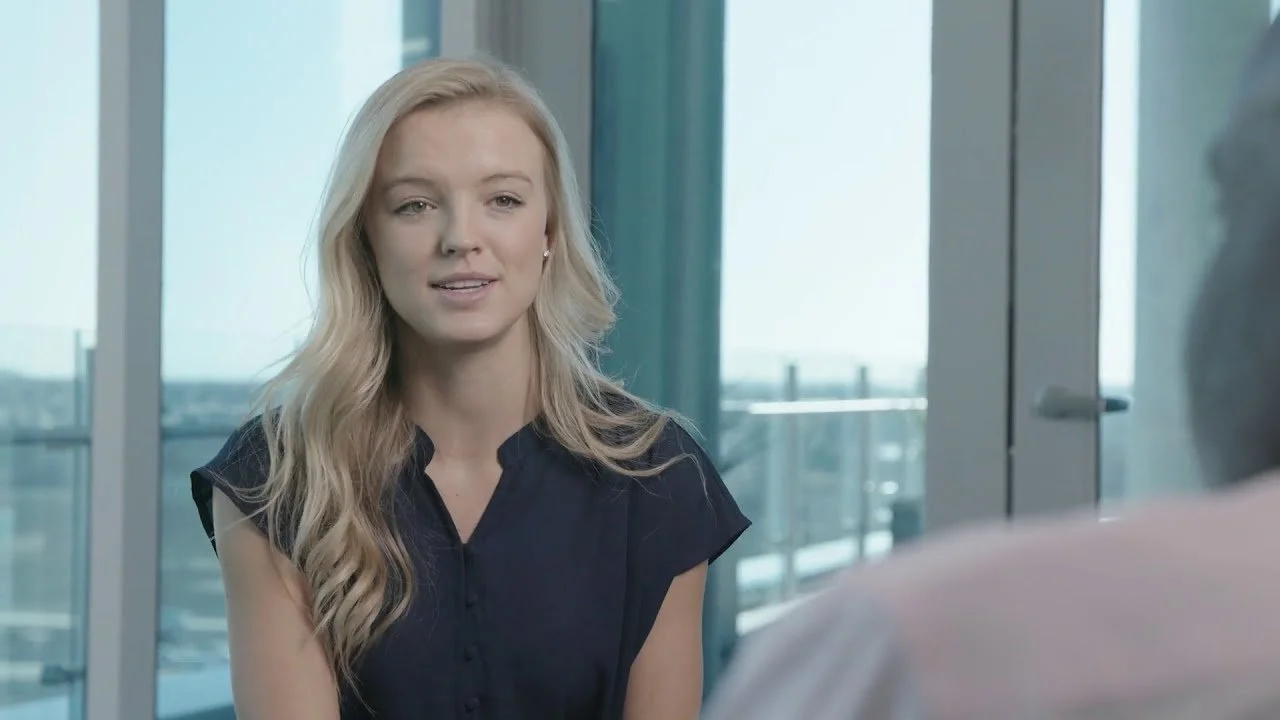 A woman with long blonde hair wearing a dark blue blouse, sitting indoors near large windows, speaking to someone off-camera.