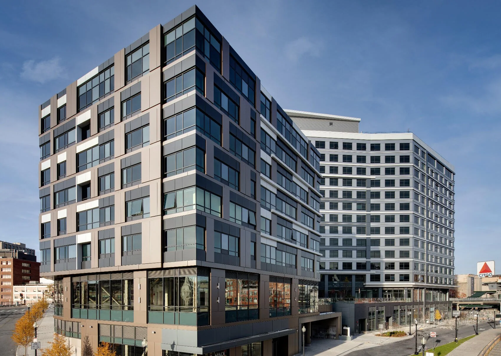 Modern multi-story buildings with glass windows and geometric design in an urban setting, with trees and clear sky in the background.