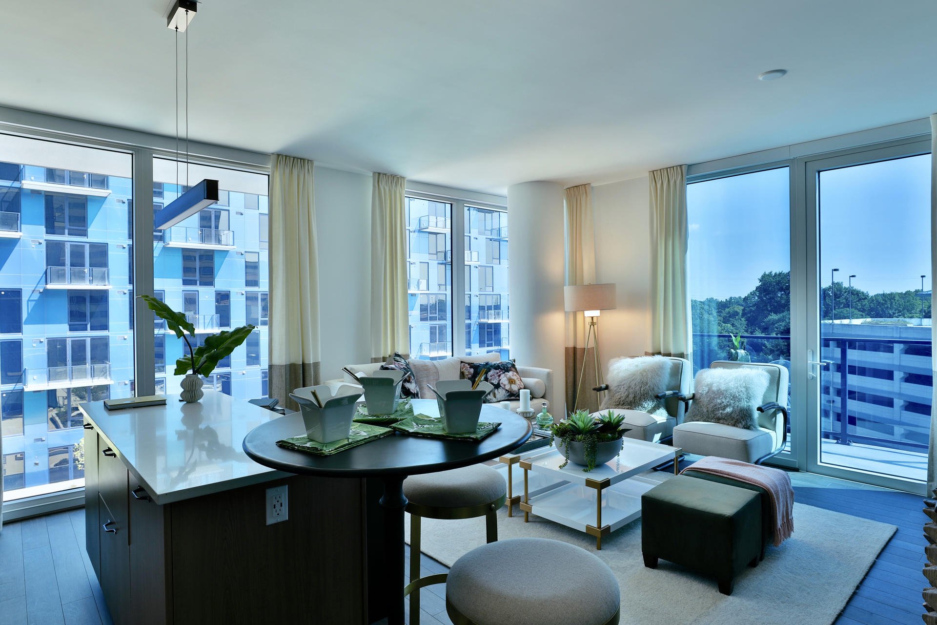 Living room with large floor-to-ceiling windows, white curtains, modern furniture, a white sofa with floral pillows, a white chair with fluffy pillow, coffee table with plants, and a cityscape view outside.