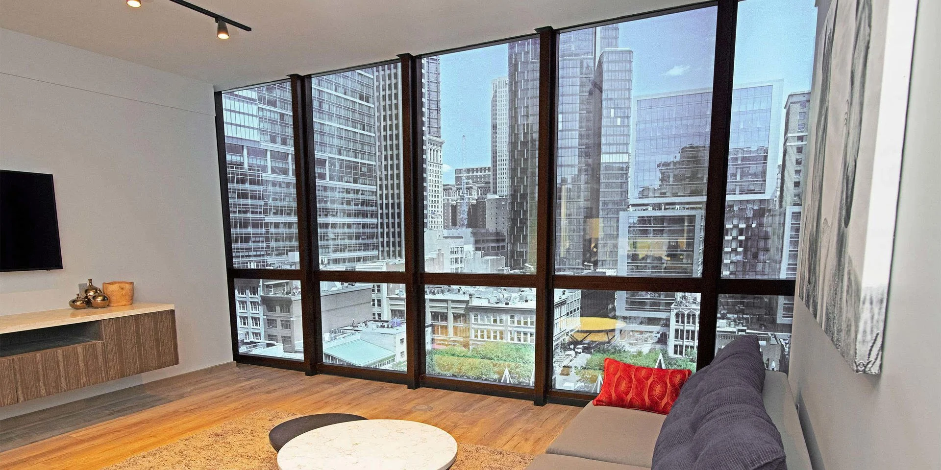Modern living room with large floor-to-ceiling windows overlooking a city skyline of tall office buildings, with a gray sofa, a red cushion, a round coffee table, and a wall-mounted TV.