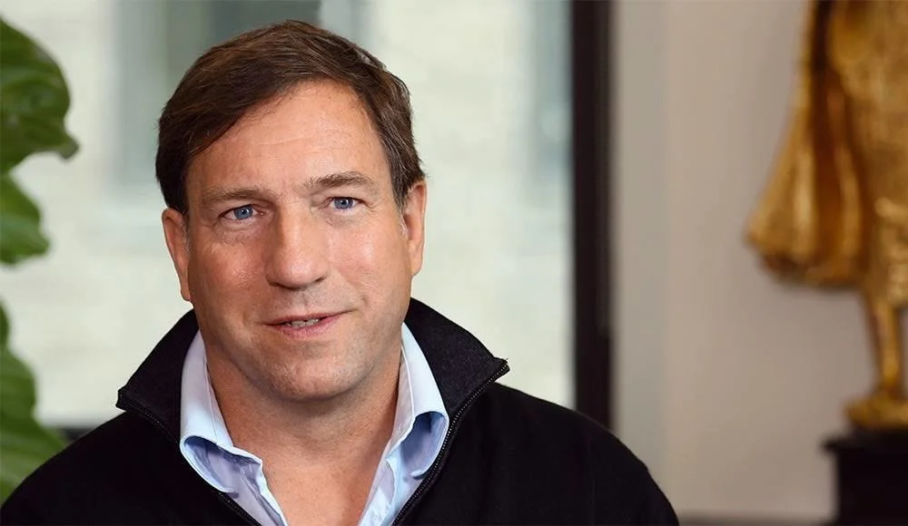 Close-up of a man with brown hair and blue eyes, wearing a light blue shirt and black sweater, standing indoors with a blurred background that includes a large green plant and a golden statue.