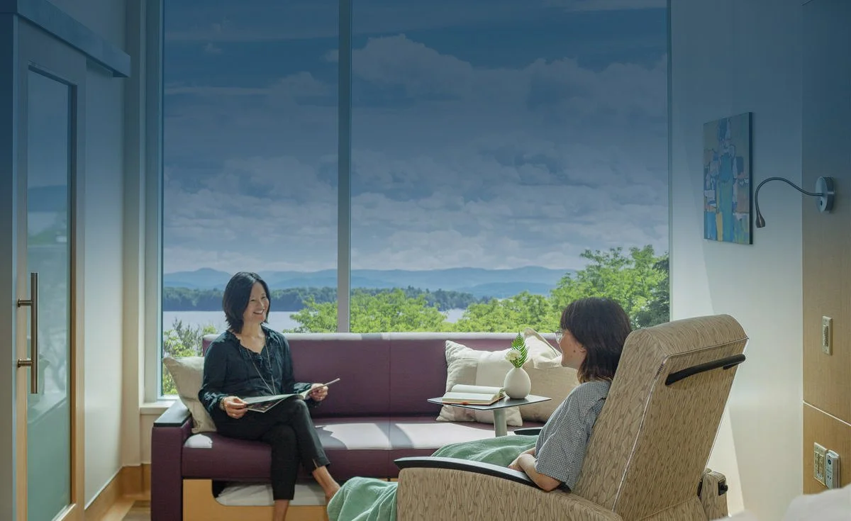 Two women sitting in a bright room with large windows overlooking a lake and mountains, having a conversation. One woman is on a sofa holding a magazine, and the other is in a recliner with a book on her lap. There is a small table with a plant between them.