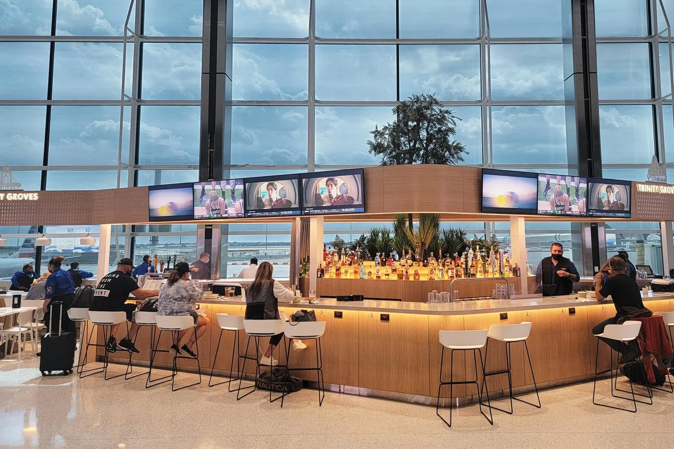 An airport bar with a curved wooden counter, bar stools, and tables with seated travelers. Multiple TV screens above the bar display a woman and a sunset. Large windows reveal an airport tarmac and planes outside. Inside, people are sitting, drinking, and using laptops, with a bartender behind the counter.