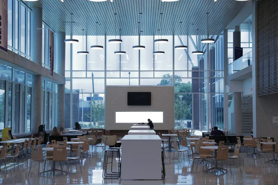 Modern, spacious indoor cafe or lounge with high ceiling, large glass windows, and minimalistic design. Few people are seated at tables, and a person is standing at the counter or serving area.