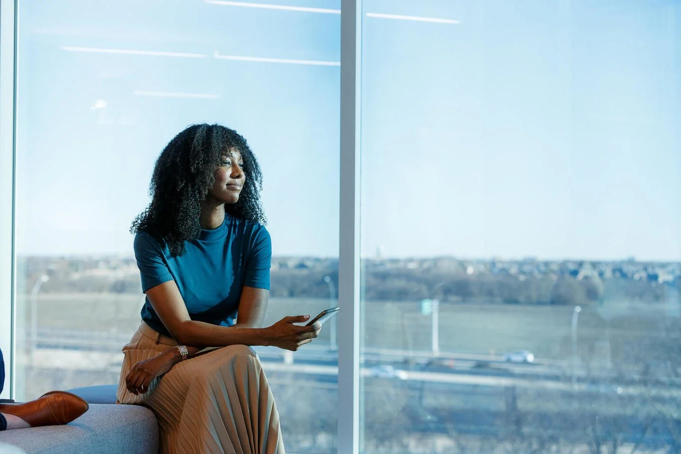 A woman sitting by a large window, holding a smartphone, looking outside at a sunny day with trees and fields in the distance.