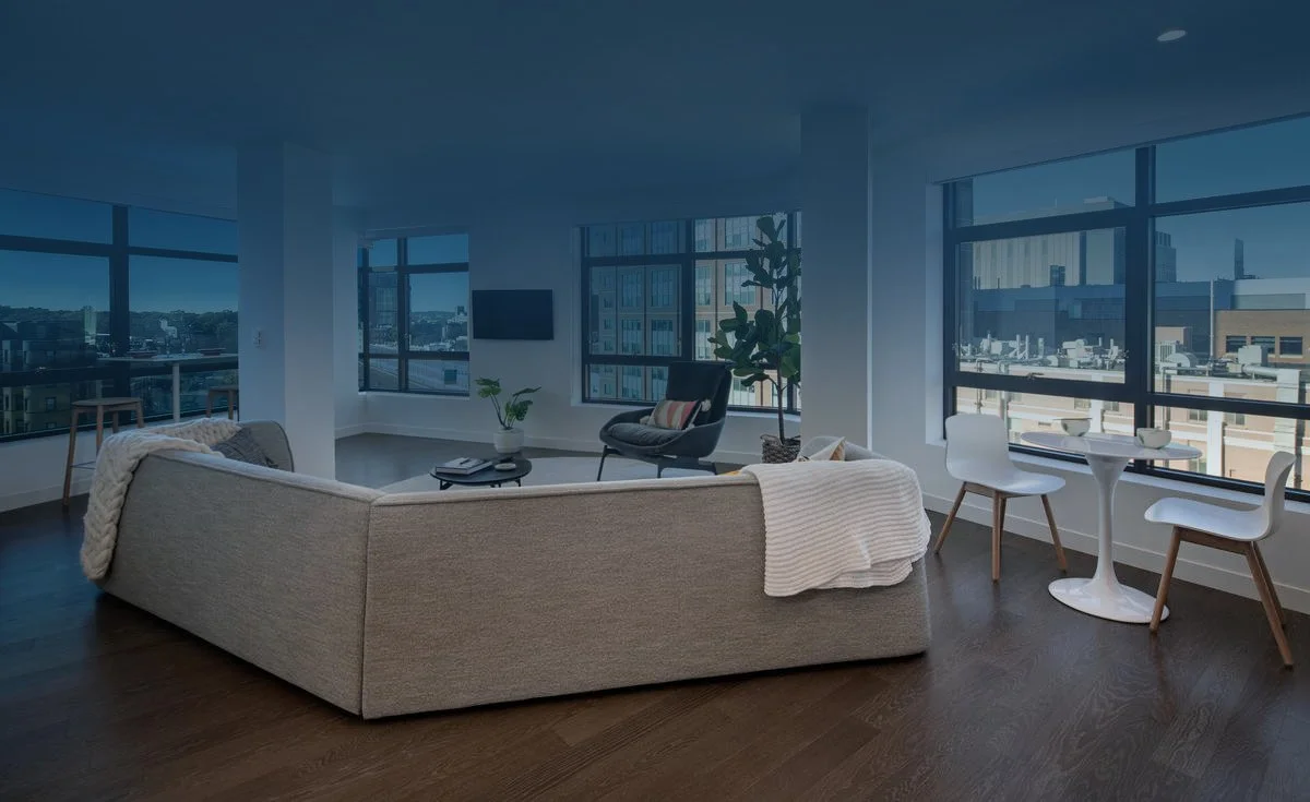 Modern living room with large windows, sectional sofa, armchair, small round dining table with two chairs, and city view.
