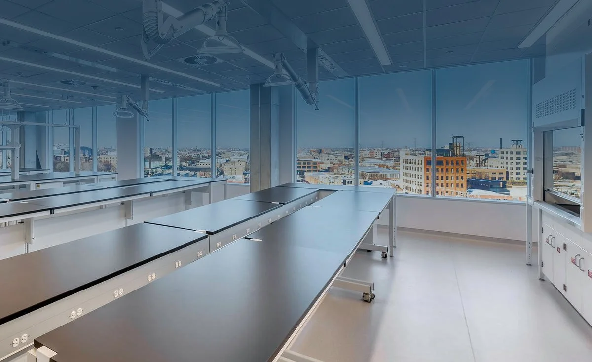 Empty medical or laboratory room with large windows and cityscape view, featuring tables, ceiling lights, and medical equipment.