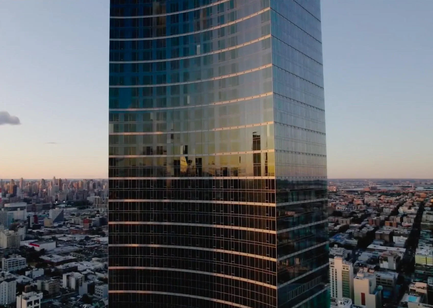 Tall glass skyscraper reflecting buildings and sky during sunset, with cityscape below.