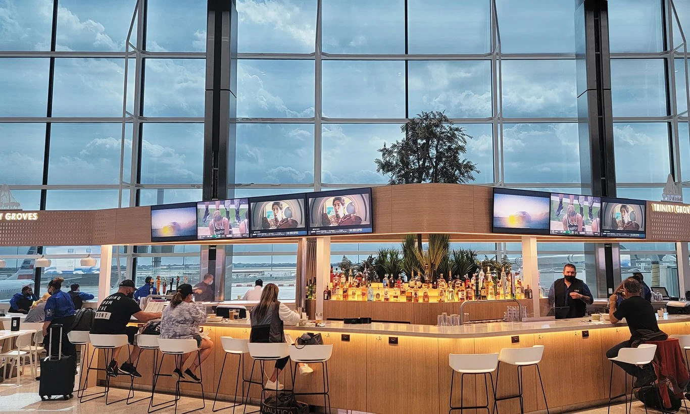 View of an airport bar area with people sitting at the counter, bartender serving drinks, large windows with cloudy sky outside, and multiple screens showing various images.