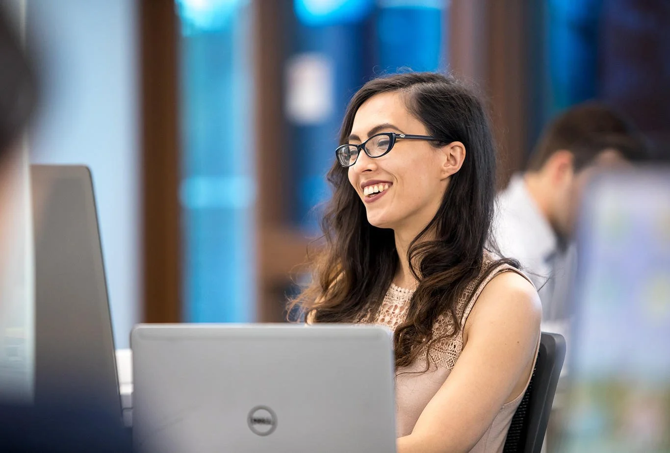 A smiling woman with dark hair and glasses working on a laptop in a modern office setting.