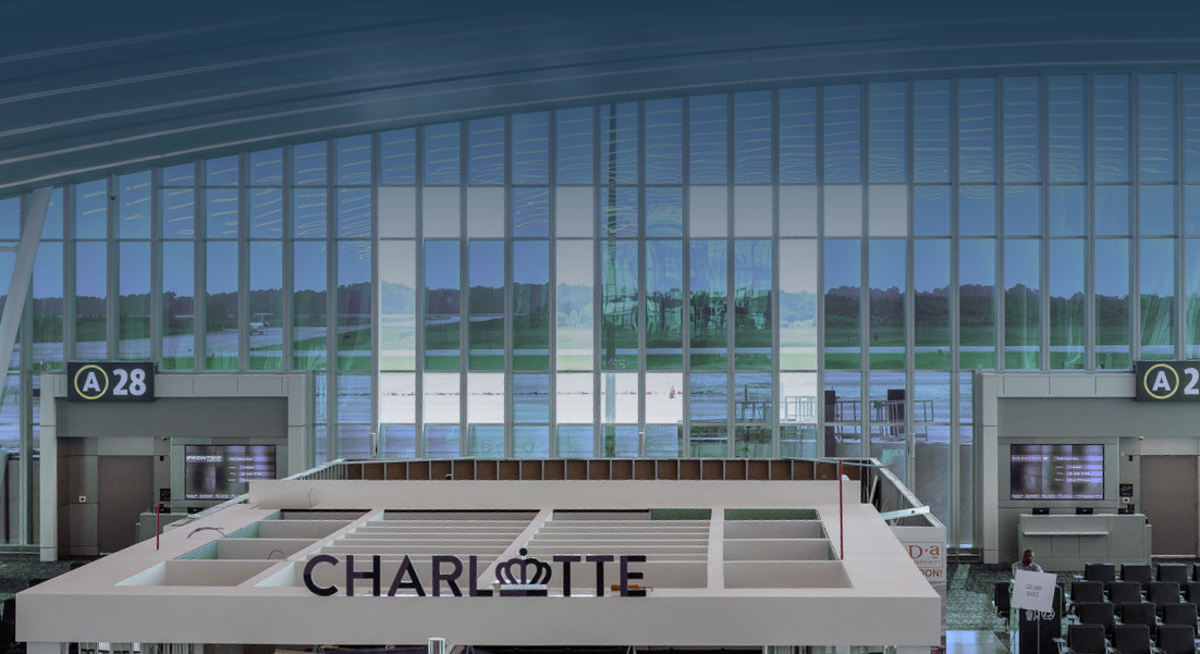 Interior of an airport terminal with large glass windows showing the runway outside, with a sign reading 'CHARLOTTE' and gates labeled A28.