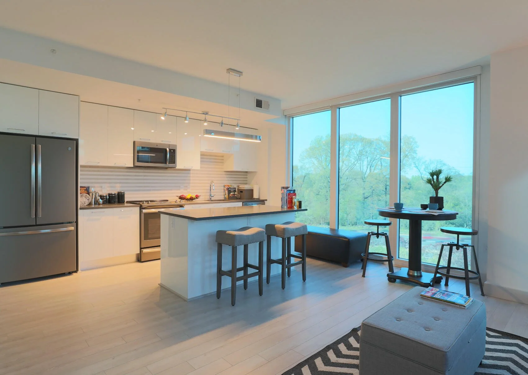 Modern kitchen with stainless steel refrigerator, oven, microwave, white cabinets, a breakfast bar with stools, and a small dining area with a round table, chairs, and a potted plant near large windows displaying trees outside.