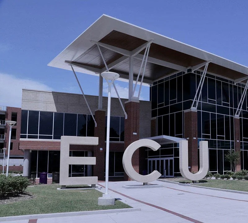 Modern building with large glass windows, a unique roof design supported by angled beams, and white letters spelling 'ECU' in front.