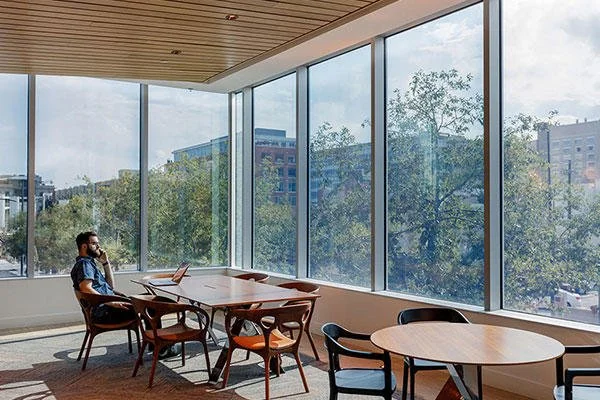 A man sitting at a table in a bright, modern office or meeting room with large floor-to-ceiling windows showing trees and city buildings outside.