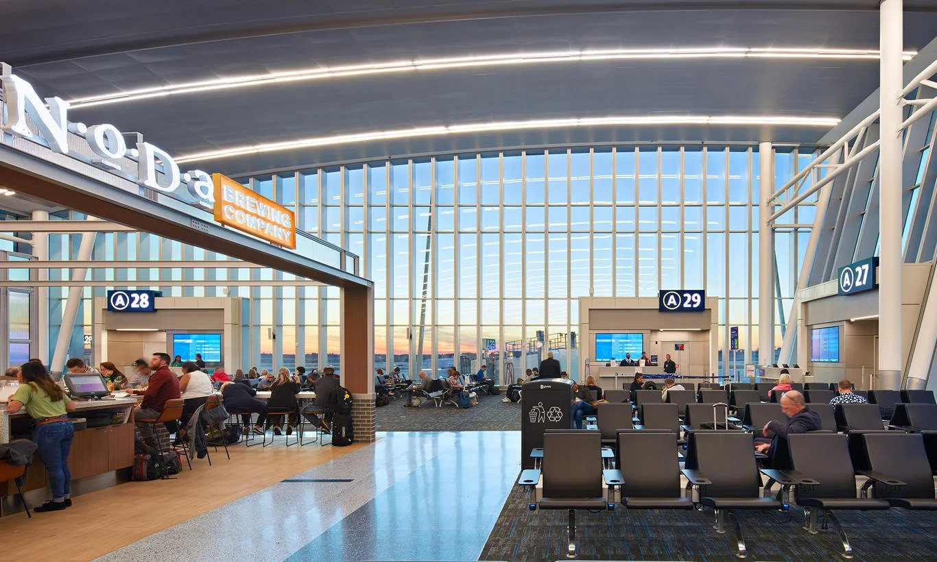 Interior of an airport with seating, check-in counters, and a view of the tarmac through large windows, during sunset.