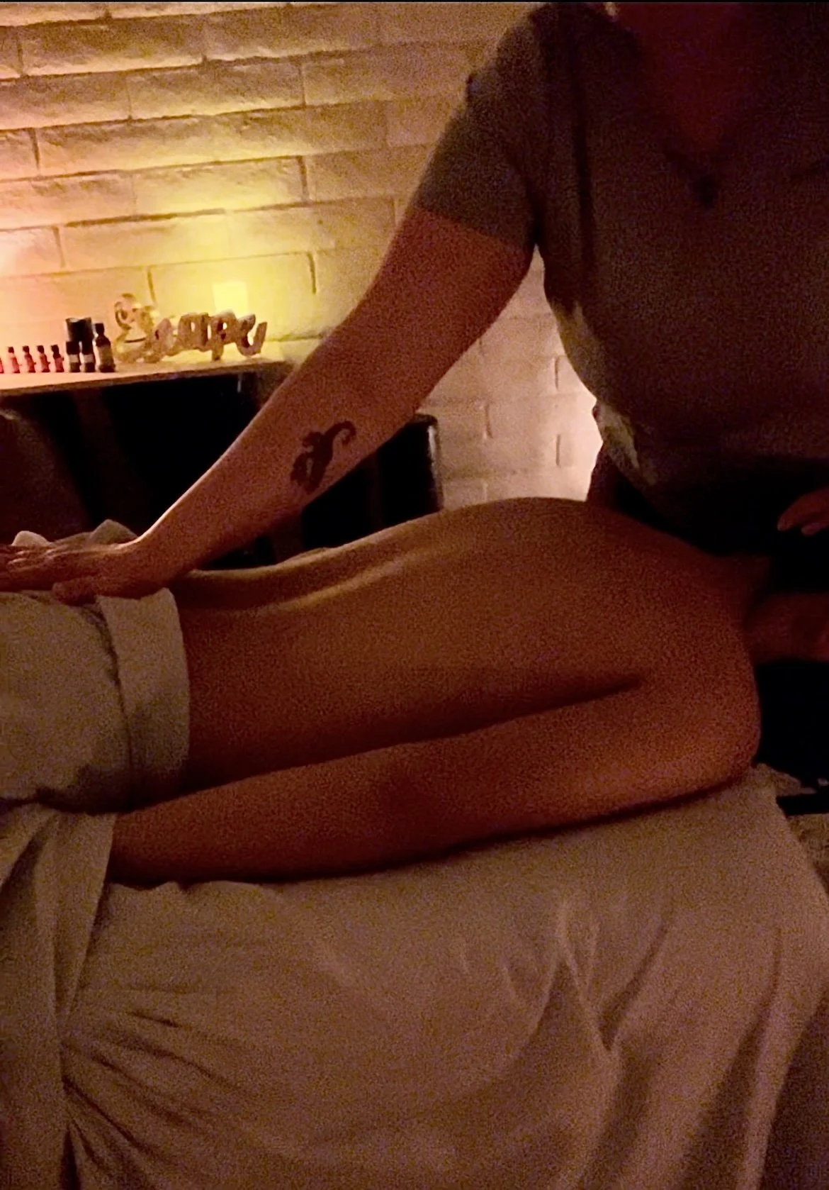 A person giving a massage to another person lying face down on a massage table in a dimly lit room with a brick wall and decorative item in the background.