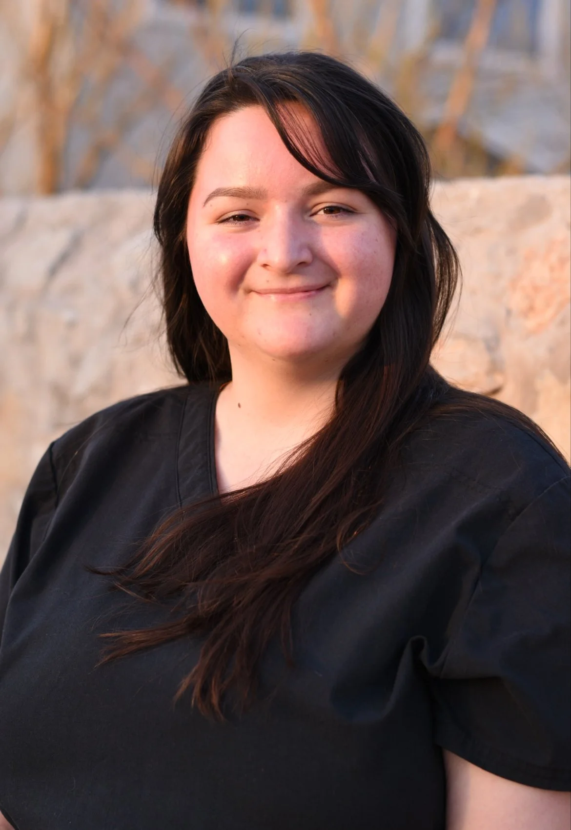 A young woman with dark hair smiling outdoors at sunset, wearing a black shirt, with a stone wall and trees in the background.