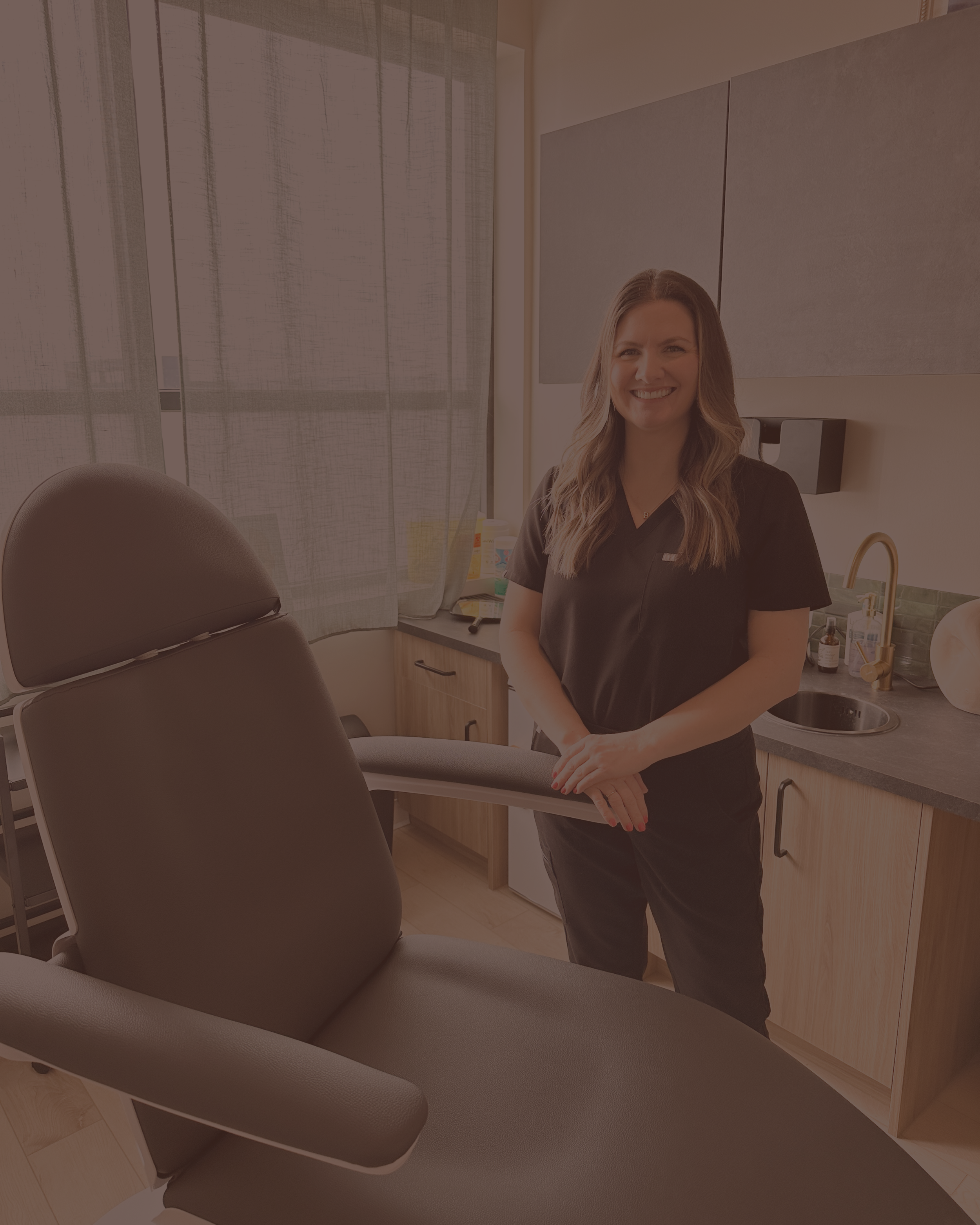 A smiling woman in black medical scrubs standing in a room with medical equipment and an examination chair.