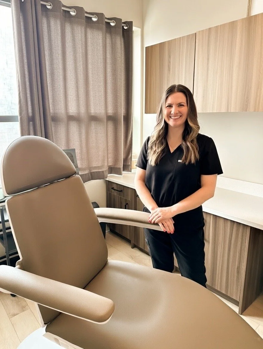 A woman in medical scrubs smiling in a consultation room with an examination chair, beige and brown cabinets, and window with brown curtains.
