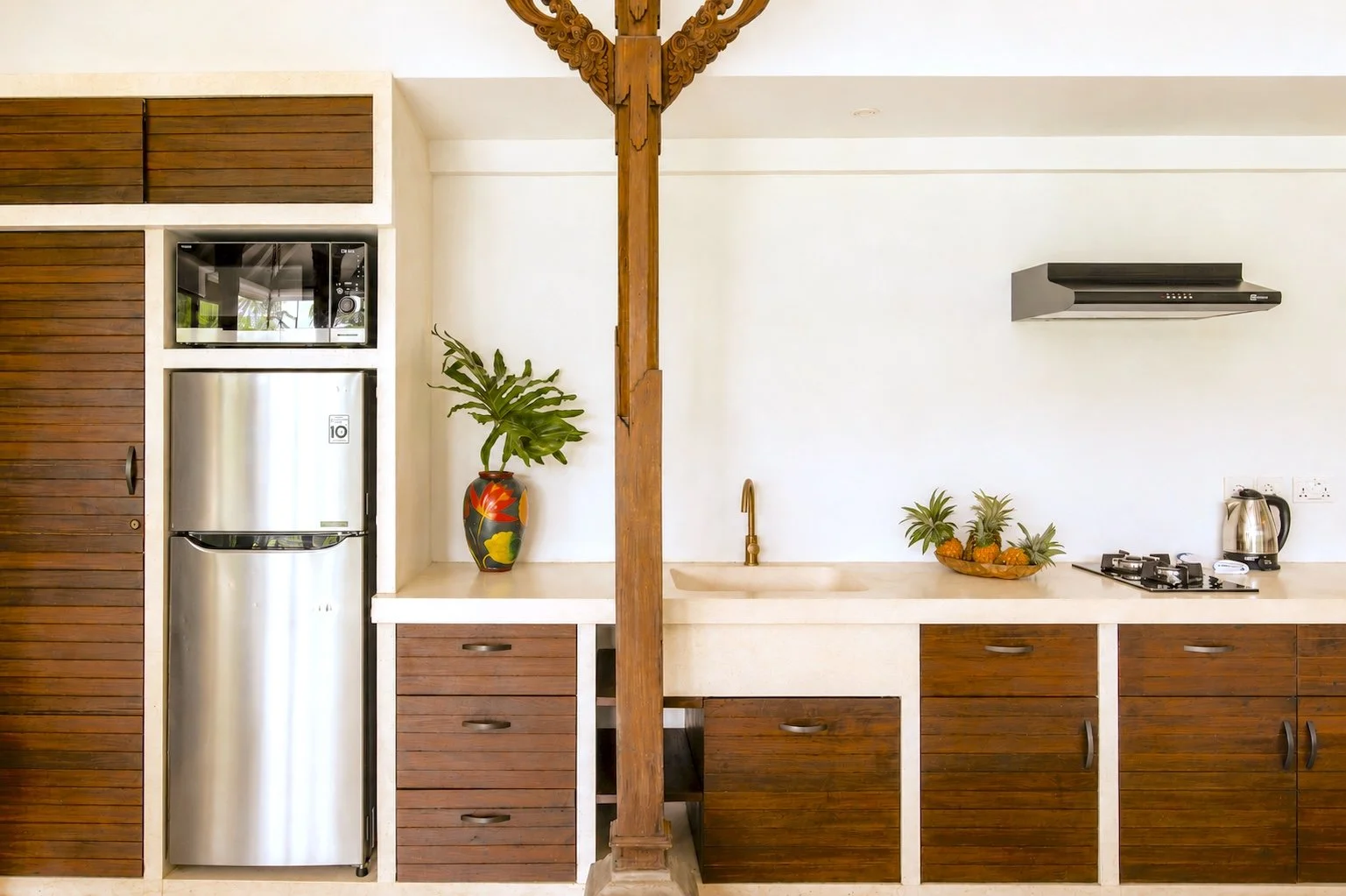 Kitchen with wooden cabinets, a stainless steel refrigerator, a microwave, a potted plant with large green leaves, a small fruit basket with pineapples, a countertop with a sink, a gas stove, and an electric kettle, with a decorative wooden column in