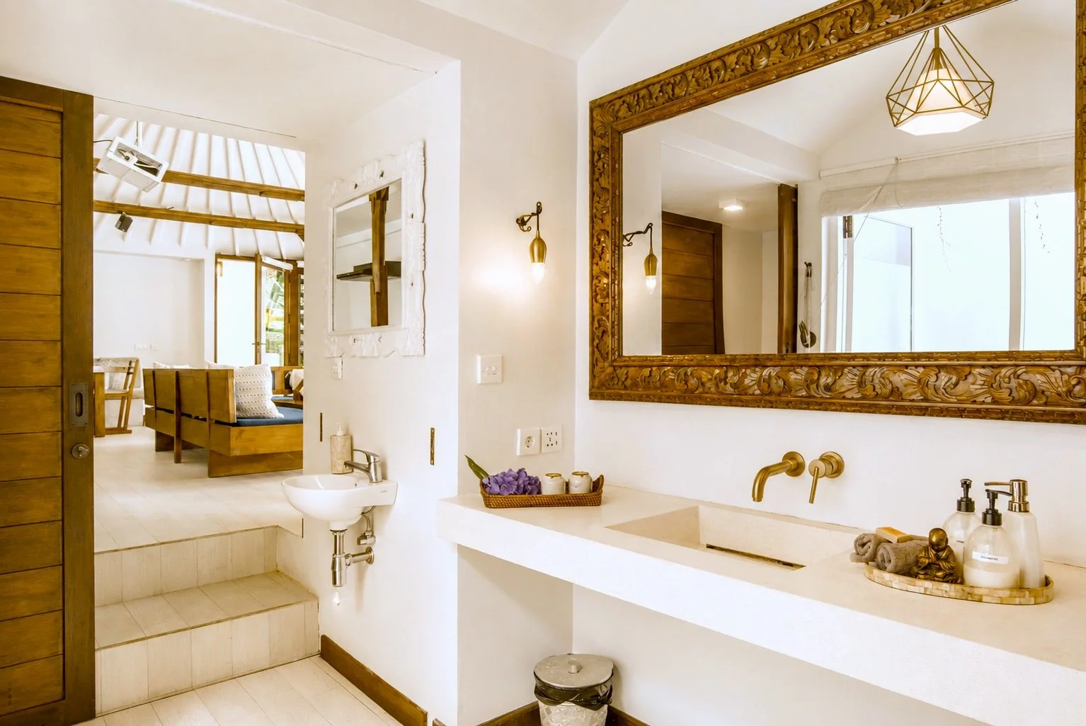 View of a modern bathroom with white walls, large mirror with ornate wooden frame, gold fixtures, and a light fixture on the ceiling. The bathroom features a beige countertop with toiletries, a sink, a small decorative tray, and a window with sheer c