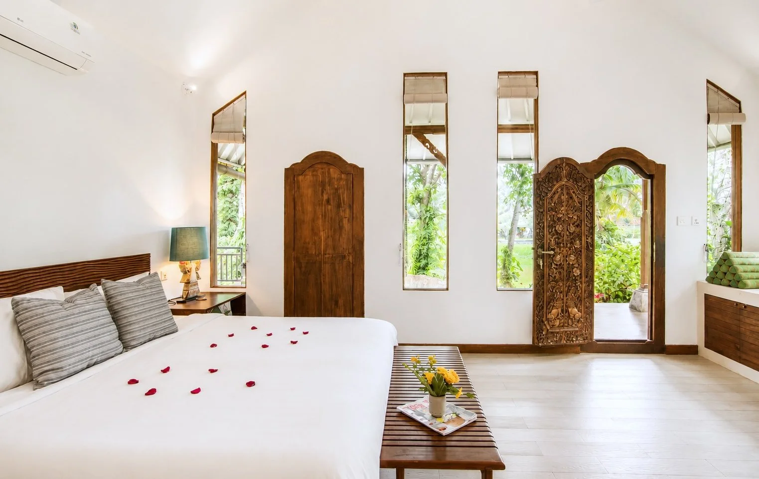 A bedroom with a large white bed adorned with rose petals, a small wooden bench with a potted plant, a nightstand with a lamp, and wooden doors and windows revealing lush greenery outside.