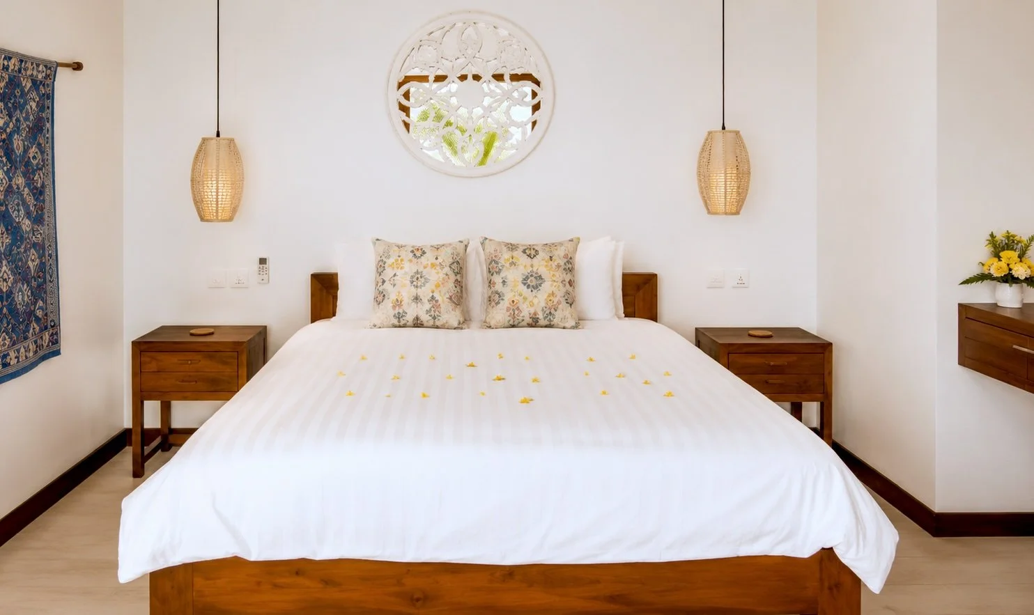 A neatly made bed with white bedding and two decorative pillows with floral patterns in a minimalist bedroom. There are two wooden nightstands on each side, hanging wicker pendant lights, a decorative round wall piece above the bed, a tapestry on the