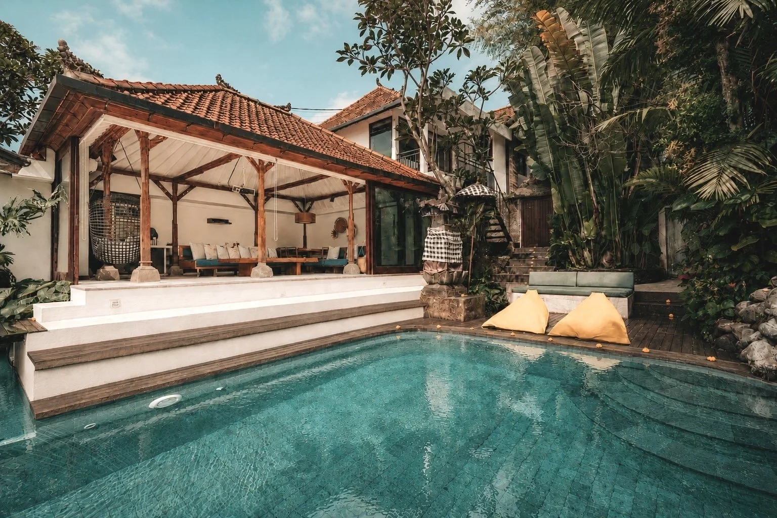 A tropical backyard with a swimming pool, wooden deck, and an open-air lounging area with cushions and hanging chairs, surrounded by lush greenery and a house with a tiled roof.