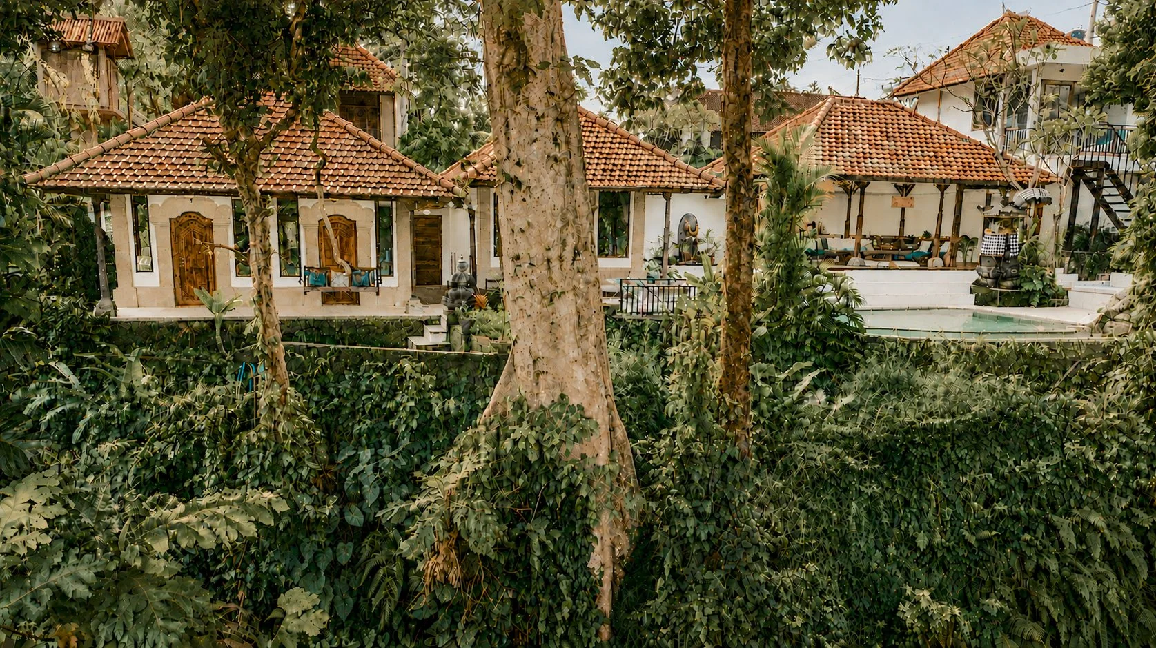 A house surrounded by lush trees with a red tile roof, outdoor seating, and a swimming pool.