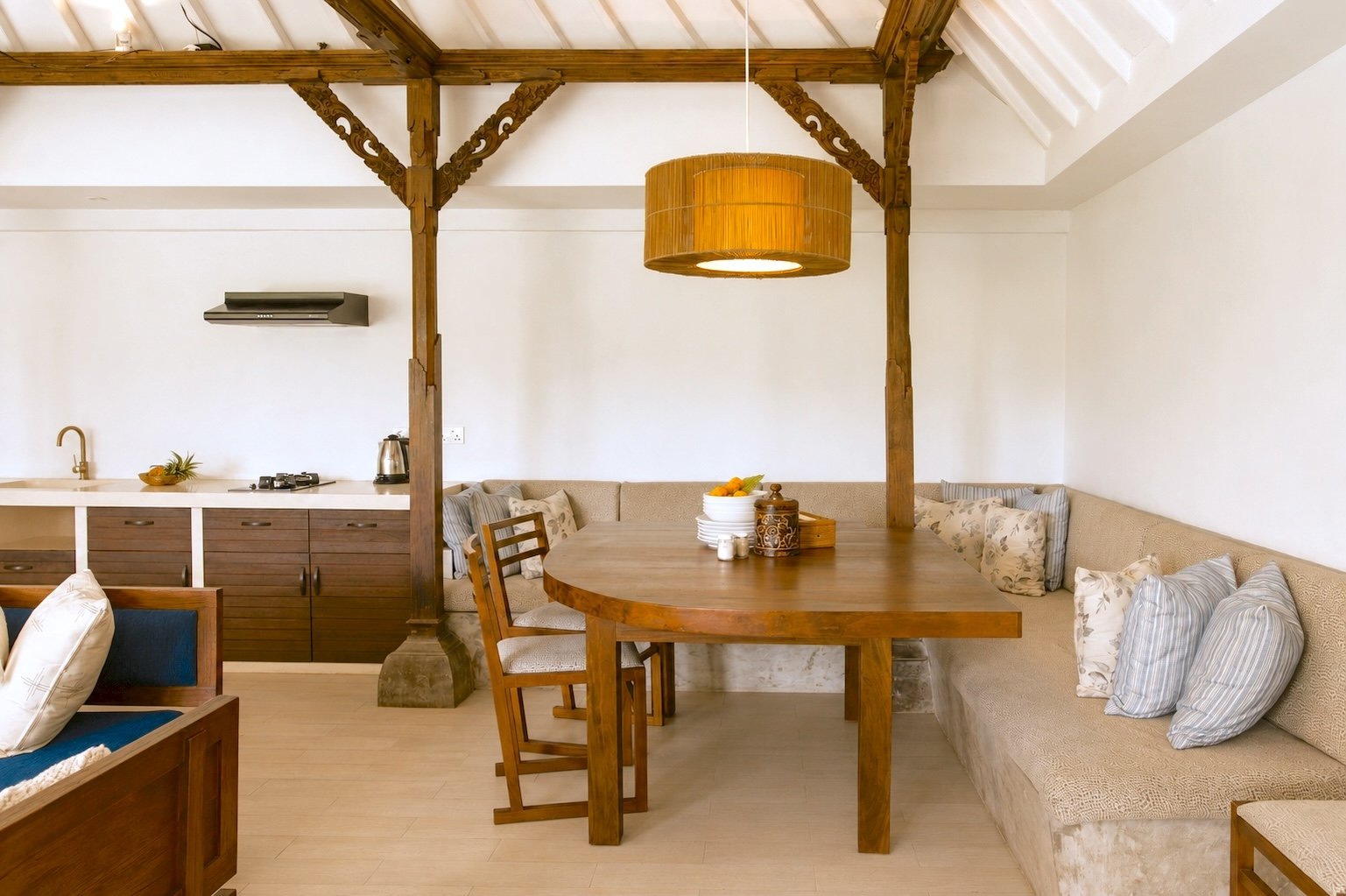 Open-concept living space with kitchen and dining area, featuring wooden beams, a large seating area with cushions, a round wooden dining table, and hanging pendant light.