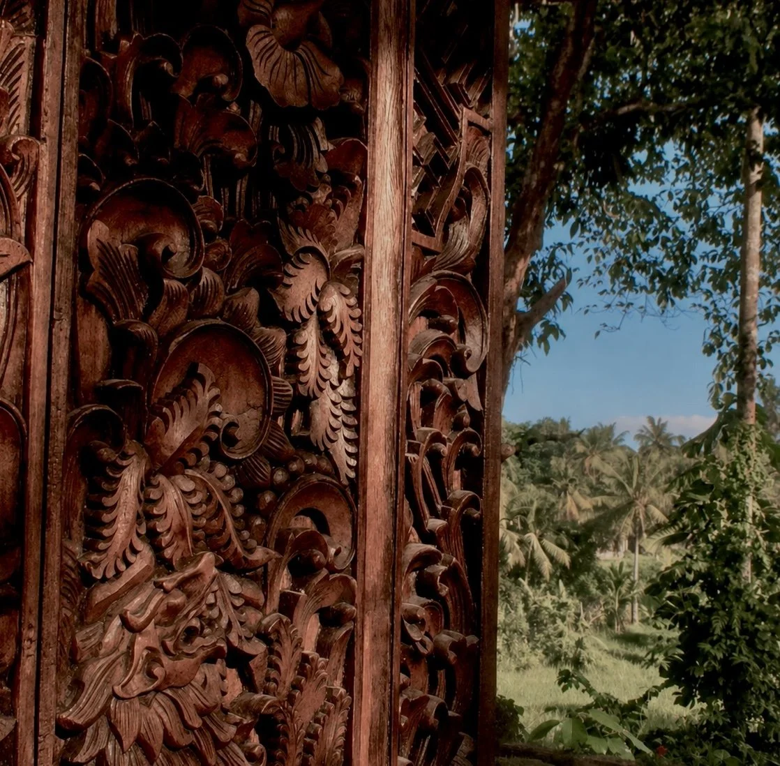 Ornately carved wooden panel with floral and leaf motifs, set against a backdrop of trees and blue sky.