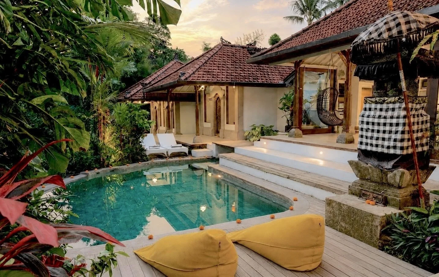 Luxury tropical villa with a small swimming pool surrounded by lush greenery, white sun loungers, and traditional Balinese architectural features at sunset.