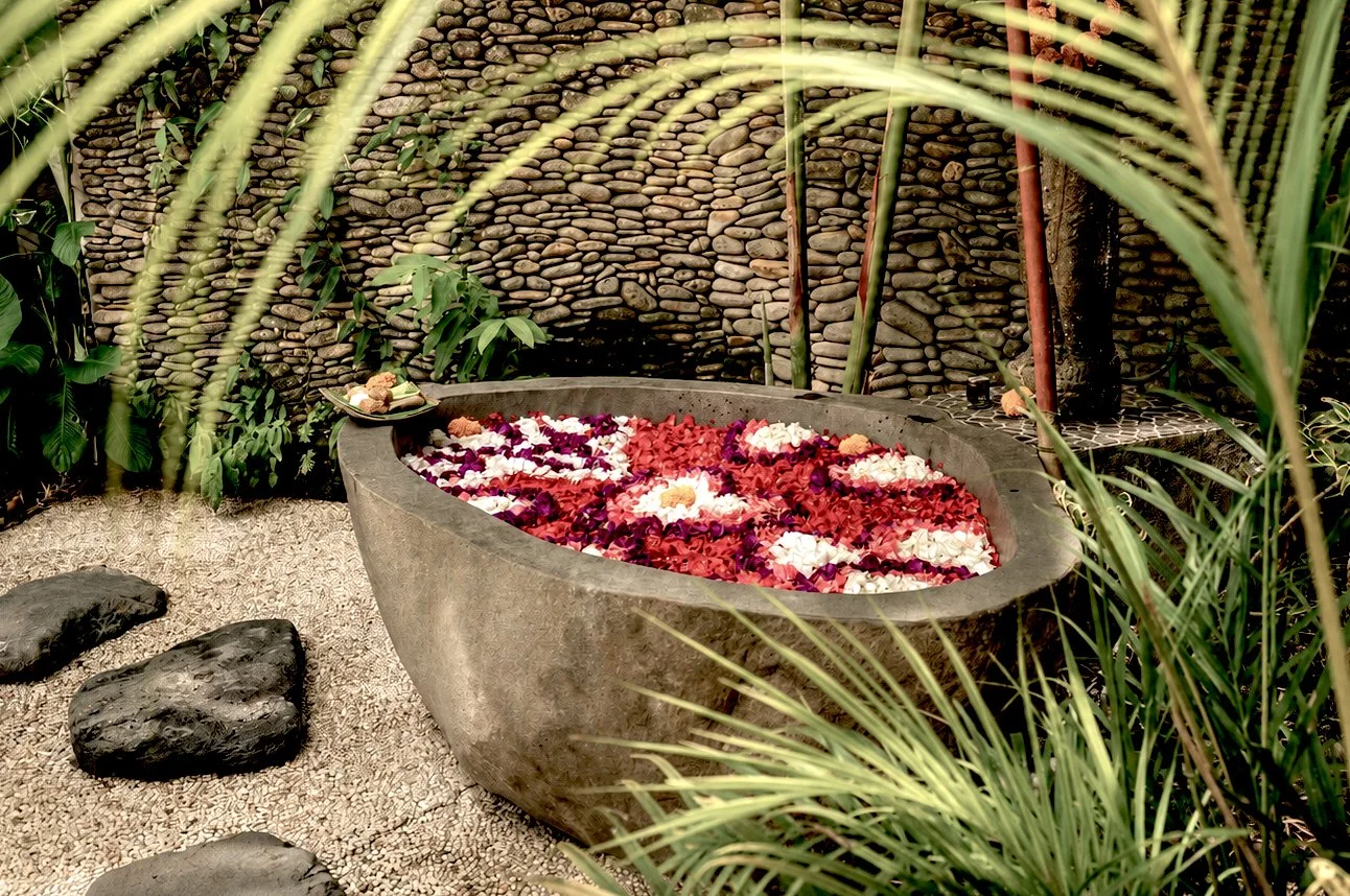 A concrete bathtub filled with floating flower petals, surrounded by green tropical plants and a stone wall, creating a tranquil garden setting.