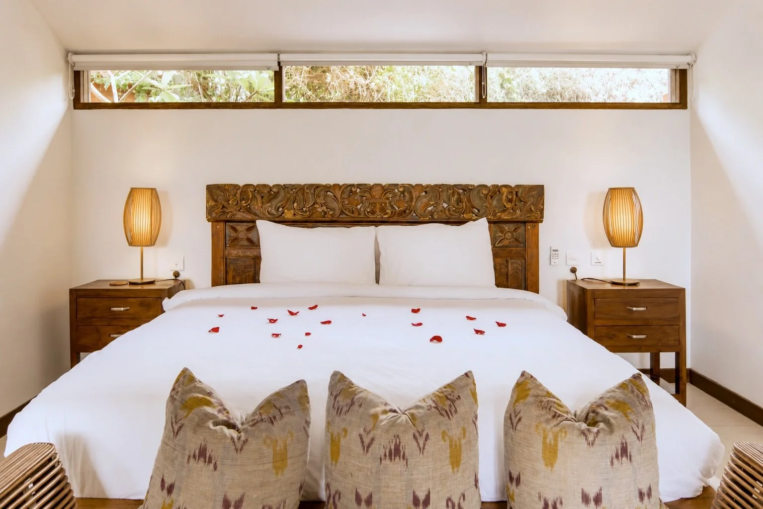 A bedroom with a large bed, decorative carved wooden headboard, white bedding with rose petals, two bedside tables with lamps, and contemporary chairs with patterned cushions at the foot of the bed. Windows above let in natural light.