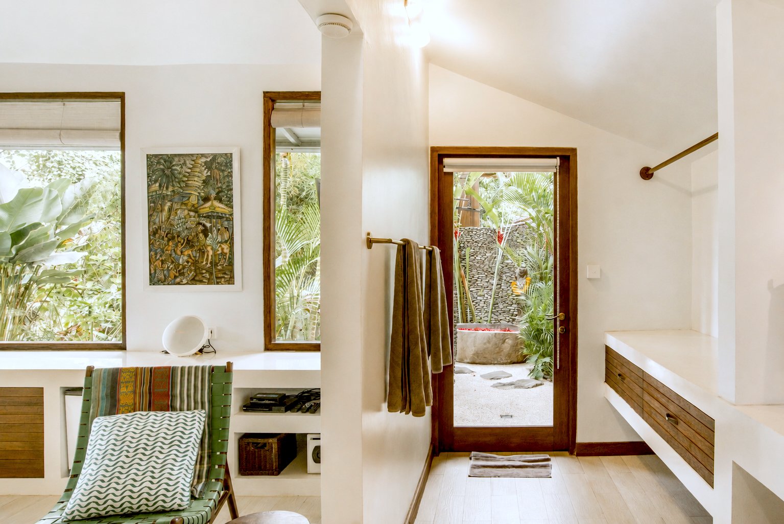 Interior view of a modern room with a door leading to an outdoor garden space. The room has two large windows with wooden frames, a chair with a cushion, a colorful piece of artwork on the wall, and towels hanging on a bar.
