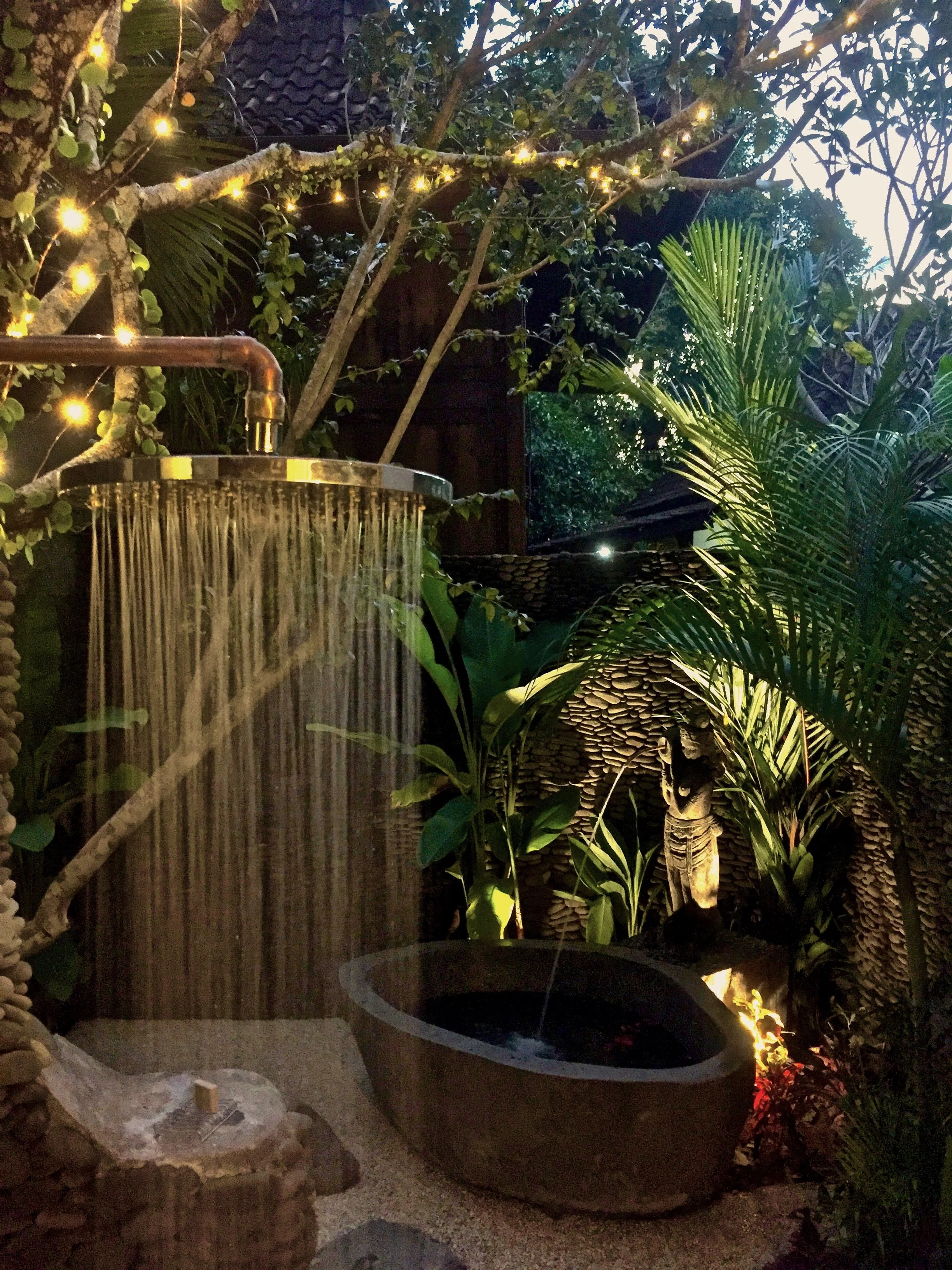 A garden scene during evening time with a waterfall feature, lush green plants, palm trees, a brick wall, and string lights illuminating the area.