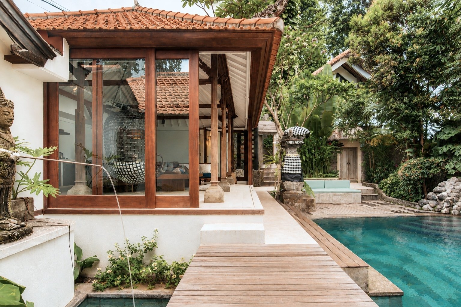 A tropical outdoor space featuring a swimming pool, a wooden deck, and a glass enclosed room with wooden supports and a tiled roof, surrounded by lush green tropical plants.