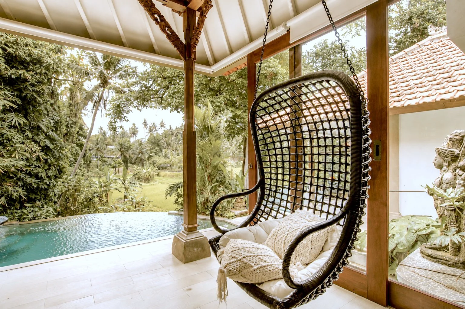 A porch with a hanging lounge chair with cushions facing a small pool and lush tropical trees and plants in the background.